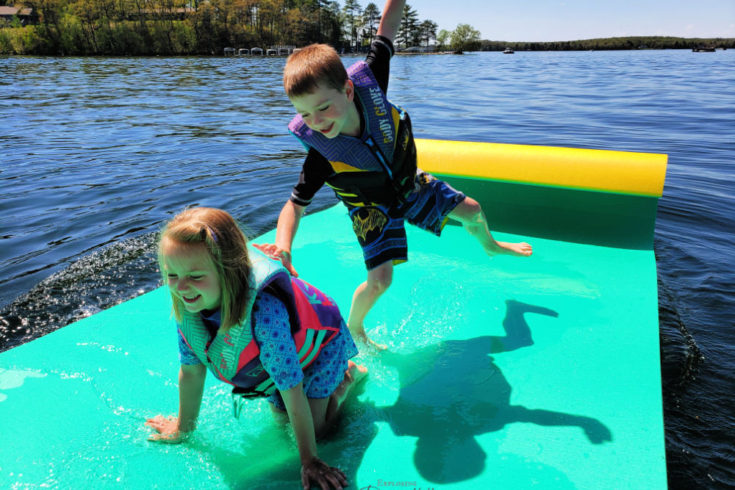 Two kids with life jackets playing on a floating mat on the water