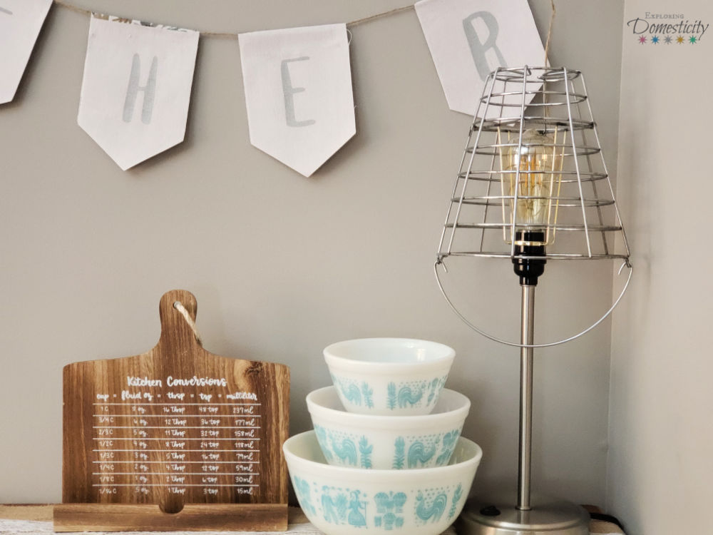 Upcycled farmhouse style lamp and basket shade with Edison bulb next to vintage Pyrex bowls and cookbook stand with conversions under "Gather" banner