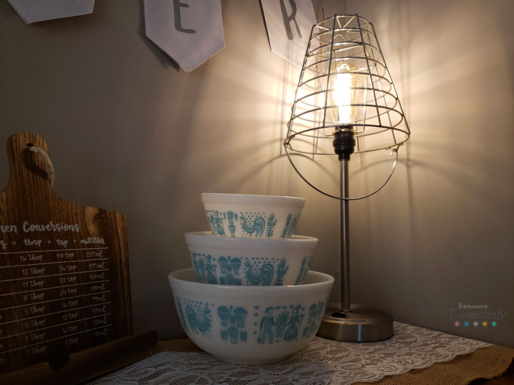 Farmhouse style upcycled lamp with wire basket shade and lit Edison bulb on burlap and lace table runner next to vintage Pyrex bowls and cookbook stand with conversions